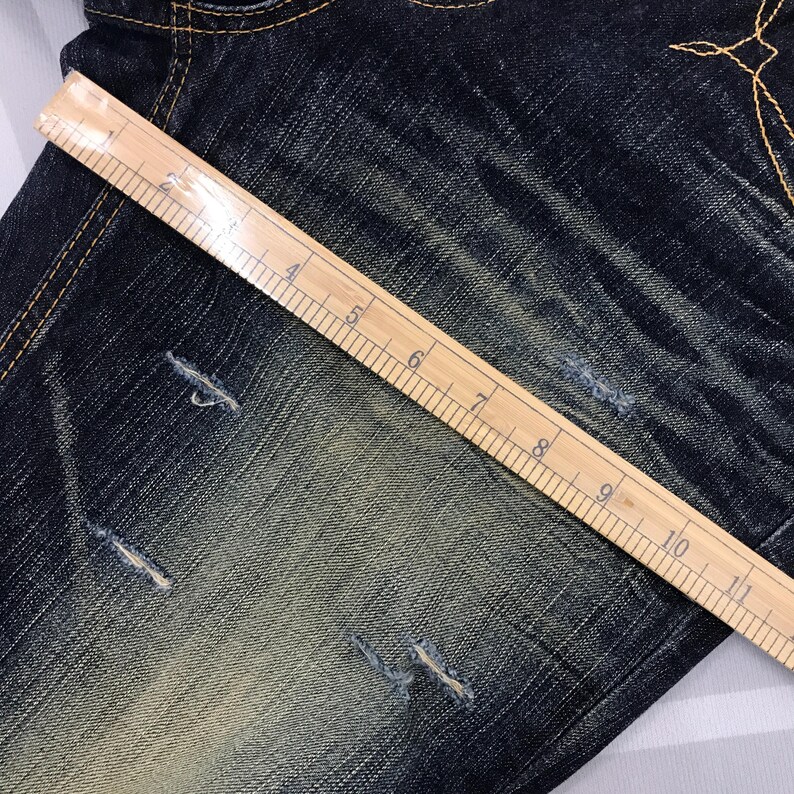 May include: Close-up of distressed blue denim jeans with a wooden ruler laid across the fabric. The ruler shows inch measurements from 1 to 10. The jeans have several small tears and faded areas.