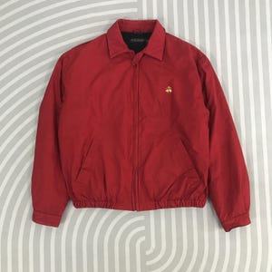 May include: A red zip-up jacket with a small embroidered logo on the chest. The jacket has a ribbed waistband and cuffs.