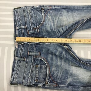 May include: Blue denim jeans with a faded wash, featuring orange stitching and classic five-pocket styling. A wooden ruler is placed across the waist, measuring approximately 16 inches across.