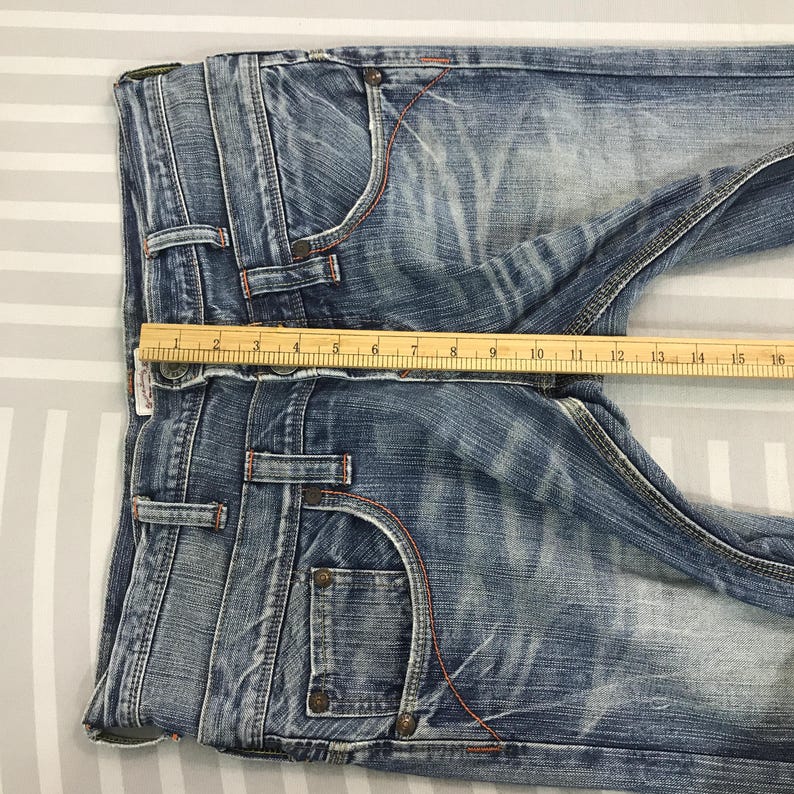 May include: Blue denim jeans with a faded wash, featuring orange stitching and classic five-pocket styling. A wooden ruler is placed across the waist, measuring approximately 16 inches across.