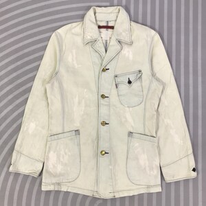 May include: A white ceramic mug with a black handle and the text "But First, Coffee" in a bold, black font. The mug is filled with a dark liquid, likely coffee, and sits on a light-colored surface.