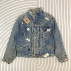 May include: A vintage blue denim jacket with multiple patches and a button-up front. The jacket features a collared neckline, two chest pockets, and two side pockets. Patches with text and graphics are sewn onto the jacket.