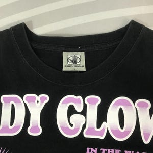 May include: Black t-shirt with the text "BODY GLOVE" in large, white and purple letters. The shirt has a crew neck and a small tag with the "BODY GLOVE" logo. The shirt is a size L.