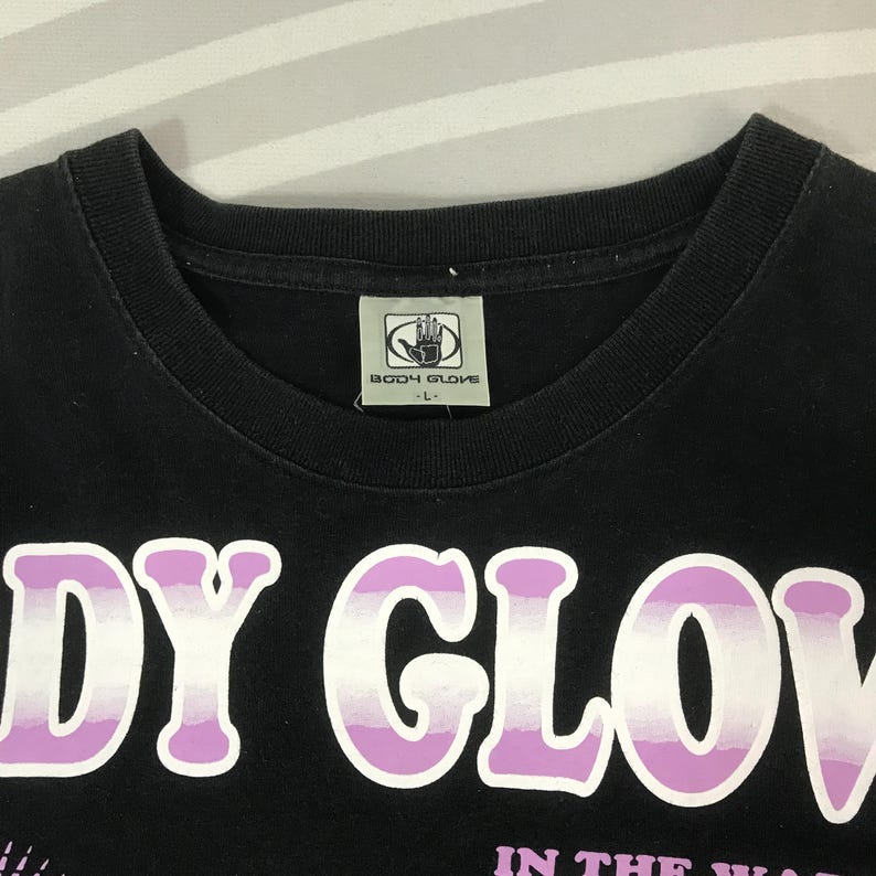 May include: Black t-shirt with the text "BODY GLOVE" in large, white and purple letters. The shirt has a crew neck and a small tag with the "BODY GLOVE" logo. The shirt is a size L.