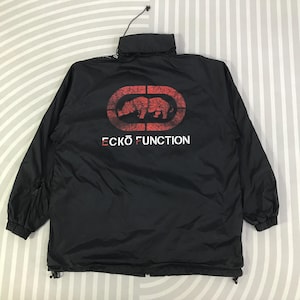 May include: A black jacket featuring a high collar with a drawstring and long sleeves with elastic cuffs. The back displays a red distressed oval graphic containing a rhinoceros silhouette above the white text ECKO FUNCTION.