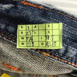 May include: Close-up of blue denim fabric with orange stitching. A yellow tag displays measurements: B 23, W 774, W 17.5, I 31.5, O 42. The denim has a textured appearance.