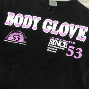 May include: Black t-shirt with the text "BODY GLOVE" in white and pink lettering. The shirt also features the number "53" and the phrase "Worldwide protection from the harsh elements."