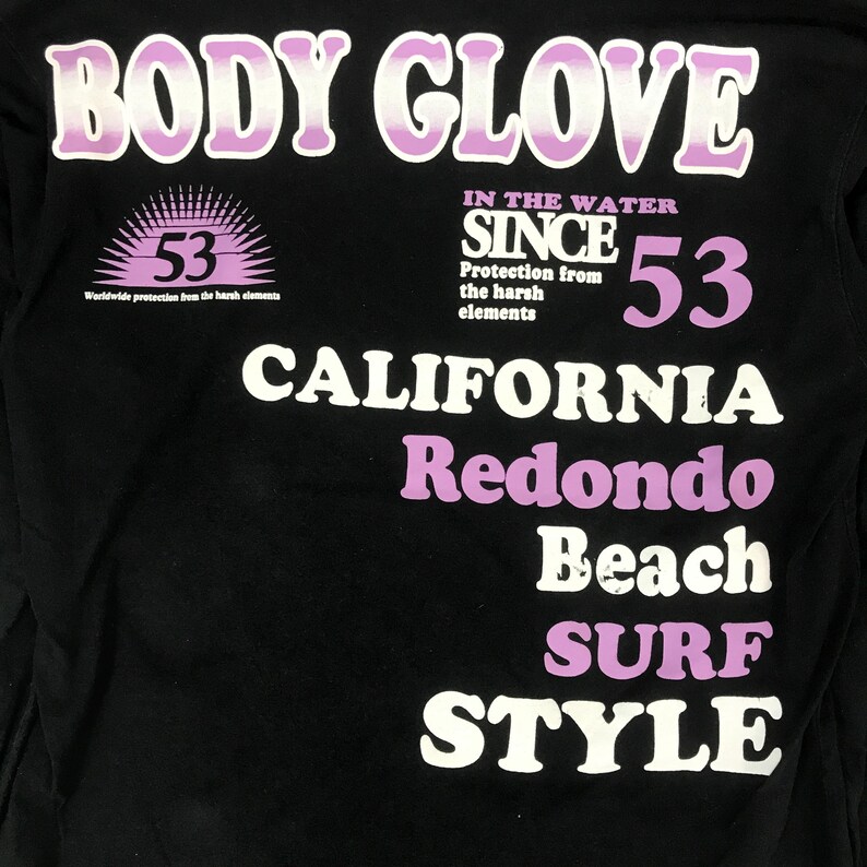 May include: Black long-sleeve shirt with the text "BODY GLOVE" in white and purple. The shirt features the text "CALIFORNIA Redondo Beach SURF STYLE" and the number "53". The shirt also has the text "IN THE WATER SINCE" and "Worldwide protection from the harsh elements".
