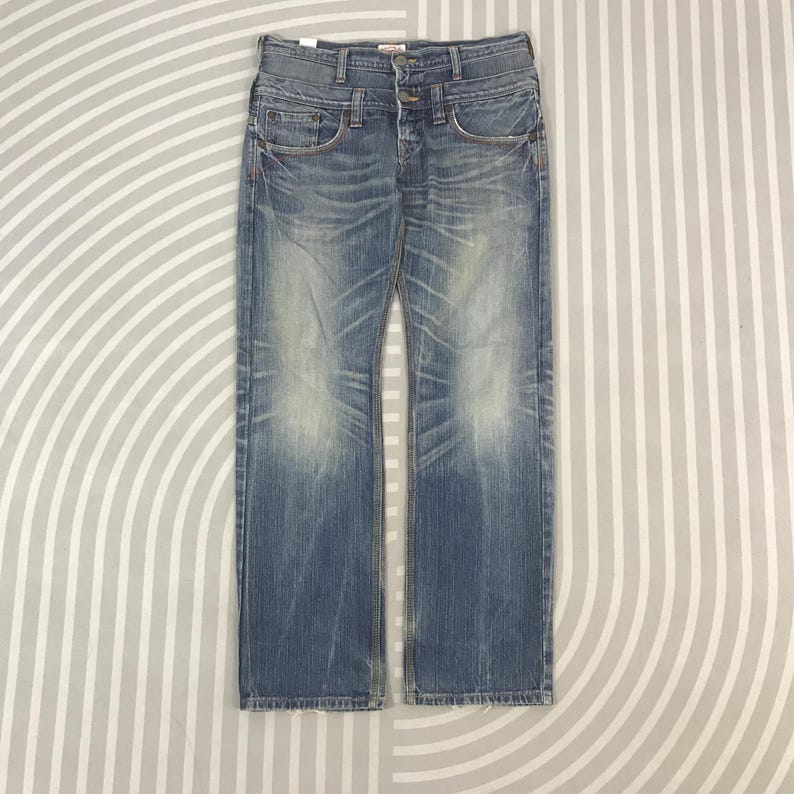 May include: Faded blue denim jeans with a straight leg cut. The jeans have a button fly, classic five-pocket design, and subtle distressing. The denim has a worn-in appearance with light fading and whiskering.