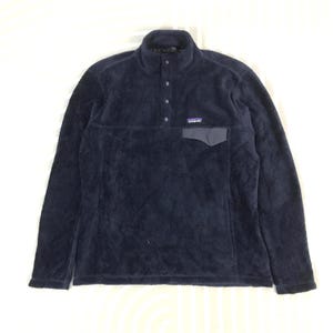 Puede incluir: Un jersey de forro polar azul marino con cuello alto y un pequeño bolsillo en el pecho. El jersey tiene cierre de botones y un pequeño logotipo de la marca en el pecho. El material parece suave y afelpado.