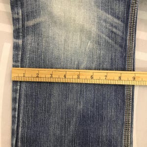 May include: Close-up of blue denim jeans with a wooden ruler measuring the width. The ruler shows measurements in inches. The jeans have visible stitching along the side seam and show signs of wear and fading.