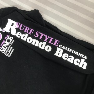 May include: Black long-sleeved shirt with white and purple text. The sleeve reads "SURF STYLE Redondo Beach CALIFORNIA" in white and purple lettering. The shirt also has text that reads "SINCE" and "Protection from the harsh elements".
