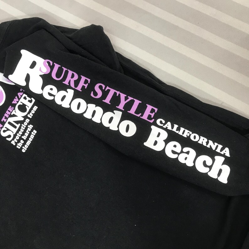 May include: Black long-sleeved shirt with white and purple text. The sleeve reads "SURF STYLE Redondo Beach CALIFORNIA" in white and purple lettering. The shirt also has text that reads "SINCE" and "Protection from the harsh elements".