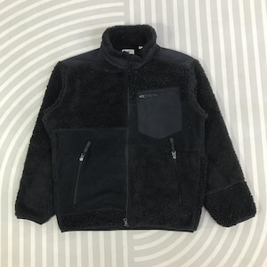May include: A black full-zip jacket featuring a high collar and a combination of textured fleece and smooth fabric panels. The garment includes a zippered chest pocket on the left side and two lower zippered pockets, all with matching black pulls.
