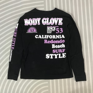 May include: Black long-sleeve shirt with "BODY GLOVE" in purple lettering. The shirt features "CALIFORNIA", "Redondo Beach", and "SURF STYLE" text. The left sleeve has "SURF STYLE CALIFORNIA" printed vertically. The shirt has a crew neck.