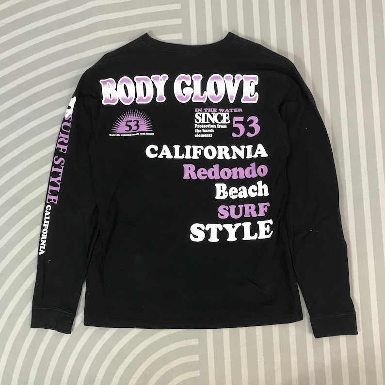 May include: Black long-sleeve shirt with "BODY GLOVE" in purple lettering. The shirt features "CALIFORNIA", "Redondo Beach", and "SURF STYLE" text. The left sleeve has "SURF STYLE CALIFORNIA" printed vertically. The shirt has a crew neck.