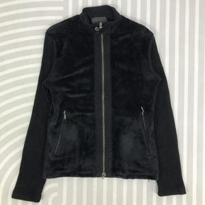 May include: Black jacket with a high collar and a full-length zipper. The jacket features a plush, velvety front panel and ribbed knit sleeves. Two zippered pockets are visible on the front. The jacket is a solid black color.