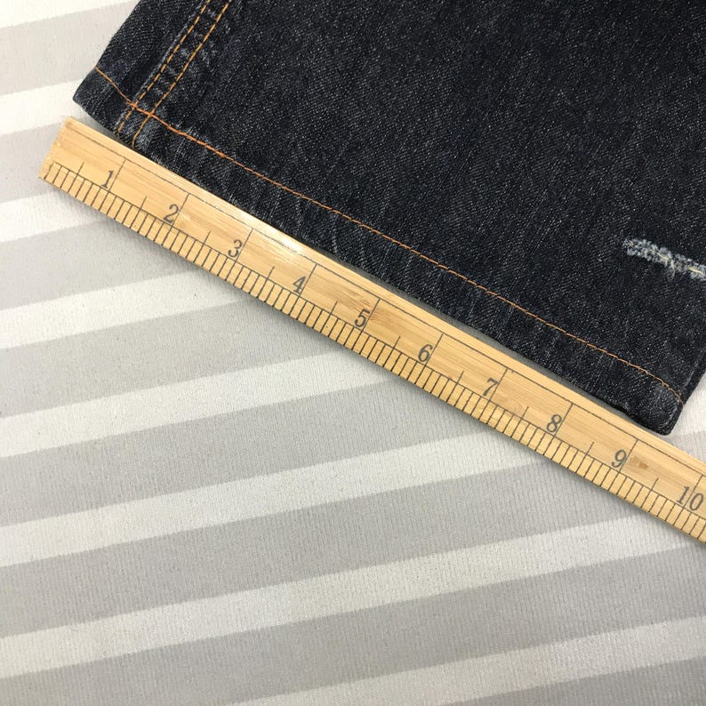 May include: Close-up of dark blue denim fabric with orange stitching. A wooden ruler, marked in inches, is placed diagonally across the fabric, showing measurements up to 10 inches. The fabric has a distressed area.