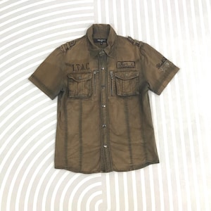 Puede incluir: Camisa marrón de manga corta con dos bolsillos en el pecho y el texto "I.T.A.C." encima de los bolsillos. La camisa tiene una parte delantera abotonada y charreteras en los hombros. El texto "Black Snake" está en la manga derecha.