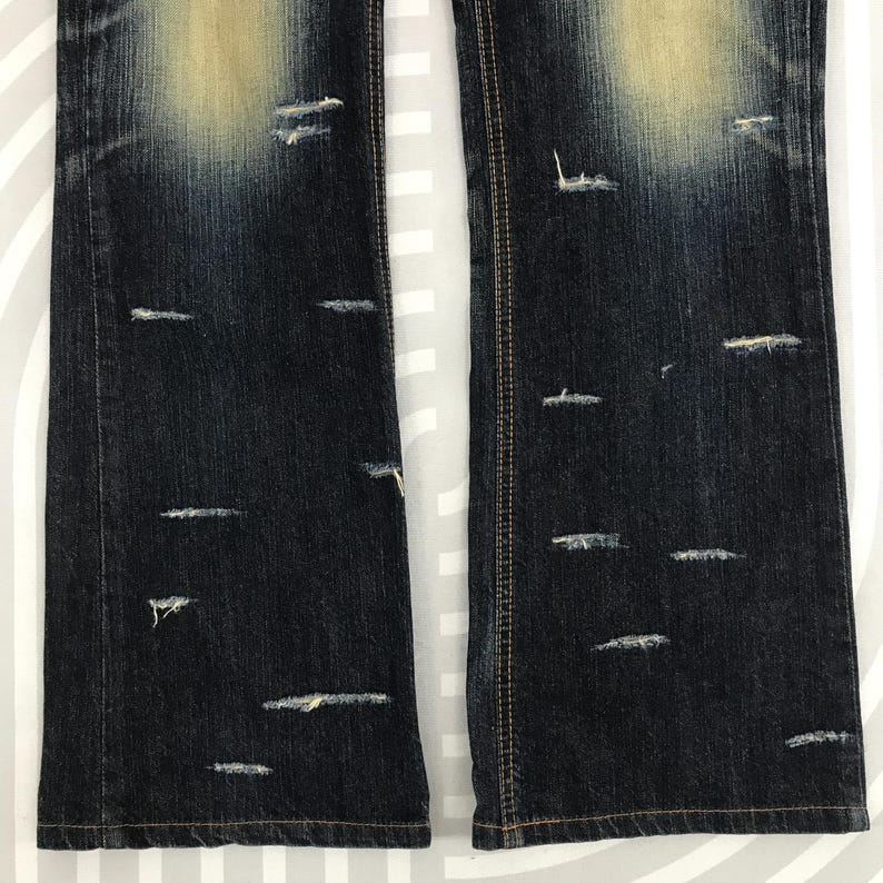 May include: Close-up of distressed dark wash denim jeans. The jeans have multiple horizontal and diagonal distressed areas. The denim has a faded appearance in the upper portion of the image. The jeans have a classic five-pocket design and a straight leg.