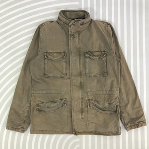 May include: A khaki-coloured jacket featuring a stand-up collar and several front pockets. The jacket has a button-up fastening and a worn look, possibly vintage. The design includes four front pockets and a zip detail near the collar. The jacket is a neutral tone.