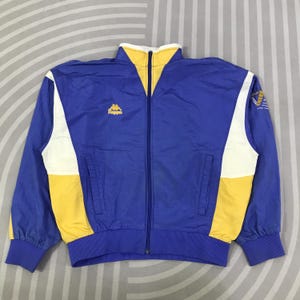 Op de afbeelding: Een blauw trainingsjack met een hoge kraag met gele accenten, een ritssluiting over de gehele lengte en witte en gele zijpanelen. Het Kappa-logo is in het geel op de borst geborduurd en er staat een grafisch ontwerp op de linkermouw.