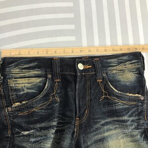 May include: Close-up of dark wash denim jeans with distressed details. Features include orange stitching, decorative pocket stitching, and a button closure. A wooden ruler is placed across the top of the jeans.