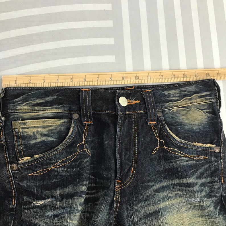 May include: Close-up of dark wash denim jeans with distressed details. Features include orange stitching, decorative pocket stitching, and a button closure. A wooden ruler is placed across the top of the jeans.