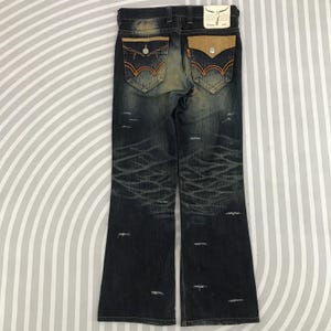 May include: Dark wash denim jeans with distressed detailing. Features include orange stitching, tan leather pocket accents, and a decorative back pocket design. The jeans have a flared leg opening.