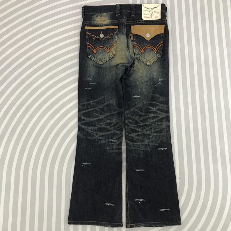 May include: Dark wash denim jeans with distressed detailing. Features include orange stitching, tan leather pocket accents, and a decorative back pocket design. The jeans have a flared leg opening.