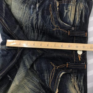 May include: Close-up of distressed blue denim jeans with faded areas and decorative stitching. The jeans have a classic five-pocket design, with a ruler showing a measurement of approximately 12 inches.