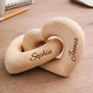May include: A light-colored wooden sculpture consisting of two interlocking heart shapes. The name Sophia is engraved in a dark script font on the left heart, and the name James is engraved in a dark script font on the right heart.