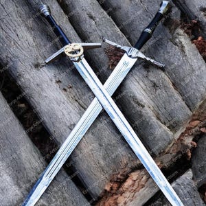 Może przedstawiać: Dwa srebrne miecze z czarnymi rękojeściami skrzyżowane na drewnianej powierzchni. Miecze mają skomplikowane detale i ostry czubek.