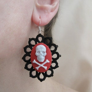 Puede incluir: Pendientes de encaje negro con un amuleto de calavera y huesos cruzados rojo y blanco.