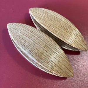 May include: Two light-colored wooden tatting shuttles with a smooth, elongated, pointed oval shape. Each shuttle features a visible natural wood grain pattern running lengthwise across the surface, with a narrow slit opening along the center.