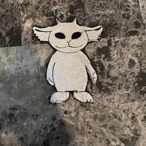 May include: A white and black embroidered patch featuring a cartoonish creature with large ears, black eyes, and small horns. The patch is in the shape of the creature and is likely designed to be ironed or sewn onto fabric.