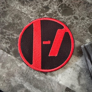 May include: A circular black patch with a red border and a red design in the center. The design features a vertical line and a horizontal line, creating a stylized symbol. The patch is likely an embroidered emblem.