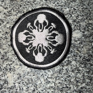 May include: A circular black patch with a white embroidered design. The design features a central shape with radiating elements, all outlined in white. The patch has a white border and a black background.