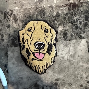 May include: Embroidered patch featuring a golden retriever dog's face. The patch has a tan and black color scheme, with a pink tongue. The dog's face is detailed with black outlines and a friendly expression. The patch is likely for clothing or accessories.
