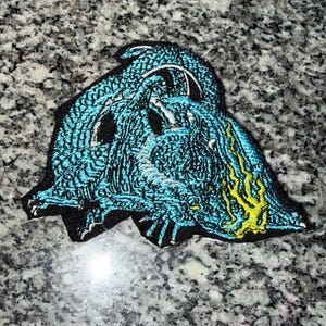 May include: A teal and black embroidered patch featuring a stylized creature design. The patch has a detailed texture with yellow accents. The design appears to be a monster or mythical figure.