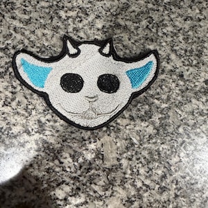 May include: A white embroidered patch featuring a stylized goat head with black eyes, small horns, and light blue accents on the ears. The patch has a black border and is likely designed for application on clothing or accessories.