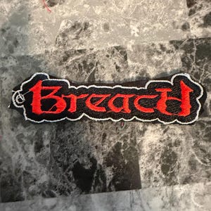 May include: Embroidered patch with the word "Breach" in red, stylized lettering. The patch has a black background with a white border. The patch is rectangular in shape.