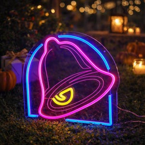 May include: A neon sign displaying the Taco Bell logo in pink, yellow, and blue. The sign is shaped like the bell and is lit with bright neon lights. The sign is placed on a grassy surface.