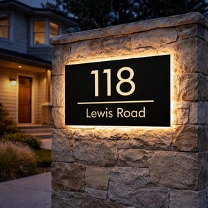 May include: Black illuminated address sign displaying the number 118 and "Lewis Road" in a modern font. The sign has a warm white glow and is set on a stone pillar. The house number is lit up at night.