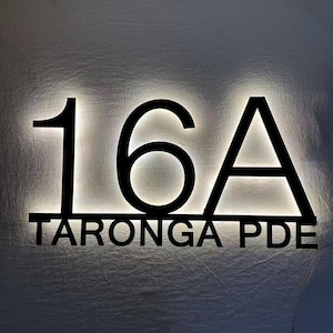 以下が含まれることがあります： サンセリフ体のフォントで「16A」という数字と「TARONGA PDE」という文字が記された黒い金属製のハウスサイン。サインは柔らかな白色の光でバックライトされており、文字と水平線の周囲にハロー効果を生み出しています。