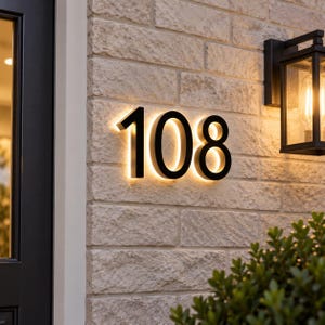 May include: A set of three-dimensional black house numbers displaying 108, mounted on a light-colored textured brick wall. The numbers feature a backlit design that casts a warm, glowing light against the brick surface, creating a halo effect.