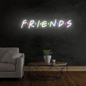 以下が含まれることがあります： 白い文字で「FRIENDS」と書かれたネオンサイン。カラフルなアクセントが付いています。このサインは、コーヒーテーブルと灰色のソファの上にある濃い灰色の壁に取り付けられています。