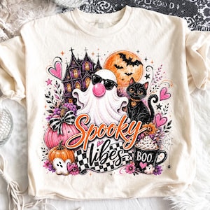 Puede incluir: Una camiseta color crema con un gráfico colorido de Halloween que muestra un fantasma con gafas de sol y una burbuja de chicle, un gato negro, una casa encantada, una calabaza y una taza con el texto BOO. El diseño incluye el texto naranja Spooky vibes.