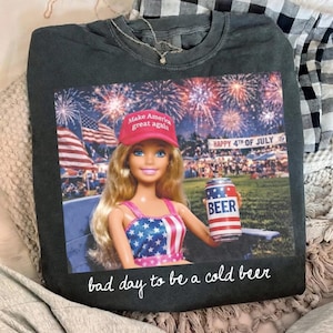 Puede incluir: Sudadera gris oscuro con un estampado de una muñeca Barbie con un sombrero rojo con el texto "Make America great again". La muñeca sostiene una lata de cerveza con un diseño patriótico, con el texto "BEER". La camiseta tiene el texto "bad day to be a cold beer."