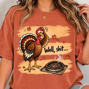 Puede incluir: Una camiseta de manga corta color óxido con un gráfico de un pavo angustiado mirando un pastel quemado en una bandeja. El pavo tiene un ala sobre la cabeza y el texto dice Well, shit... rodeado de hojas de otoño que caen.